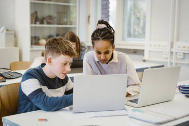 Boy sharing laptop with female friend sitting at desk in classroom - MASF38336