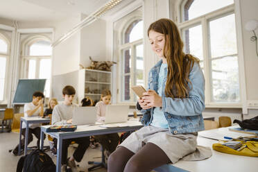 Smiling female student using smart phone while sitting at desk in classroom - MASF38339