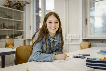 Porträt einer lächelnden Schülerin mit langen Haaren, die am Schreibtisch im Klassenzimmer sitzt - MASF38341