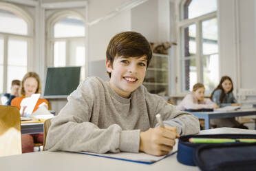 Portrait of happy schoolboy sitting at desk in classroom - MASF38342