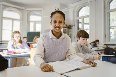 Porträt einer glücklichen Schülerin, die mit einem männlichen Freund im Klassenzimmer am Schreibtisch sitzt - MASF38343