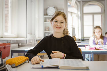 Porträt einer lächelnden Schülerin mit Brille, die mit einem Buch am Schreibtisch im Klassenzimmer sitzt - MASF38344