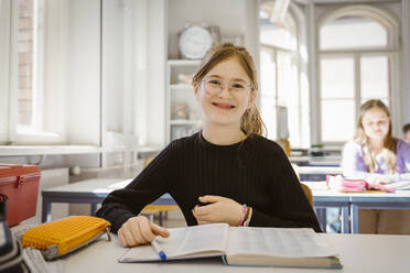 Portrait of smiling schoolgirl wearing eyeglasses sitting with book at desk in classroom - MASF38344