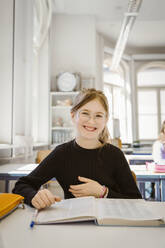Porträt einer glücklichen Schülerin mit Buch am Schreibtisch im Klassenzimmer - MASF38345