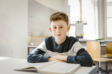 Portrait of schoolboy sitting with book at desk in classroom - MASF38347