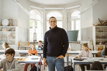 Porträt eines glücklichen männlichen Lehrers mit Hand in der Tasche, der inmitten von Schülern im Klassenzimmer steht - MASF38352