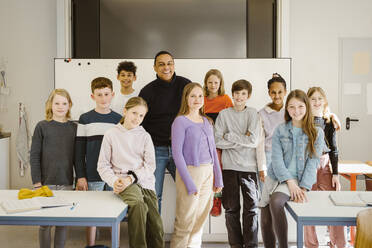 Porträt von lächelnden, gemischtrassigen Schülern, die mit einem männlichen Lehrer am Schreibtisch im Klassenzimmer stehen - MASF38353