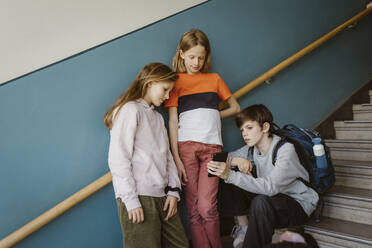 Boy sharing smart phone with female friend standing against wall on school building - MASF38356