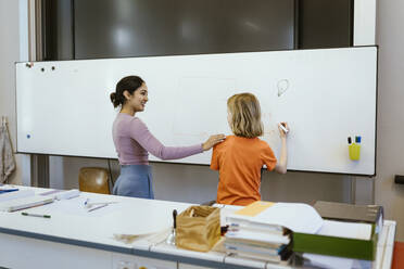Smiling female student with hand on shoulder of student writing on whiteboard in classroom - MASF38370