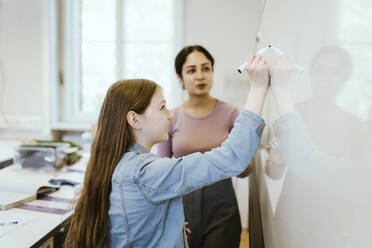 Side view of student writing on whiteboard by female teacher in classroom - MASF38372