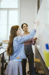 Side view of schoolgirl writing on whiteboard with felt tip pen by teacher in classroom - MASF38373