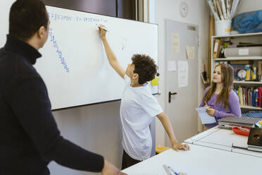 Schoolboy writing on whiteboard with teacher in classroom - MASF38376