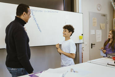 Smiling boy talking to male teacher standing near whiteboard in classroom - MASF38377