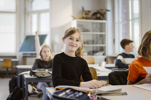 Porträt einer lächelnden Schülerin, die mit einer Freundin im Klassenzimmer am Schreibtisch sitzt - MASF38379