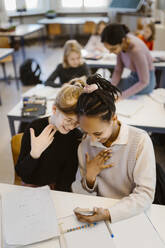 High angle view of schoolgirls gesturing while using smart phone sitting at desk in classroom - MASF38381