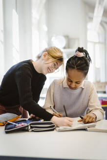 Smiling girl with female friend writing in book while studying in classroom - MASF38387