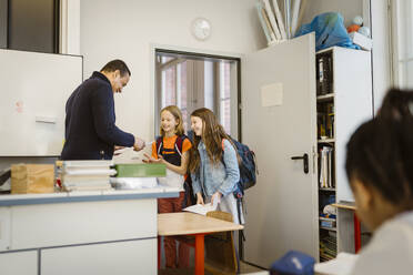 Mature male teacher giving documents to students standing in classroom - MASF38395