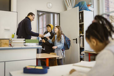 Mature male teacher giving documents to happy students standing in classroom - MASF38396