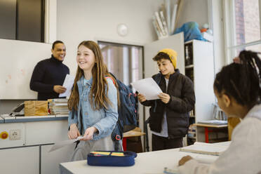 Smiling male and female students walking in classroom holding documents - MASF38397