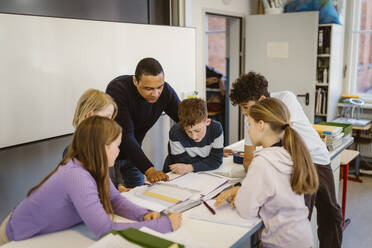 Teacher explaining male and female students leaning on desk in classroom - MASF38398