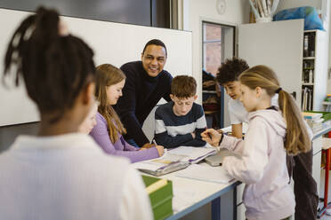 Happy teacher with multiracial students at desk in classroom - MASF38399