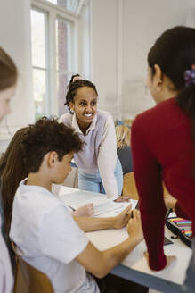 Smiling student leaning at desk with friends and teacher in classroom - MASF38404