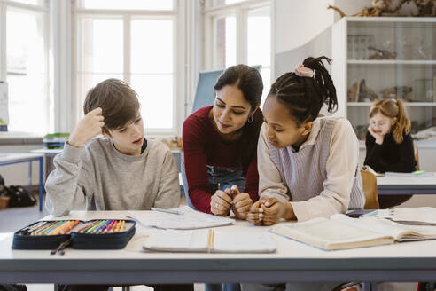 Teacher discussing with male and female students sitting at desk in classroom - MASF38411