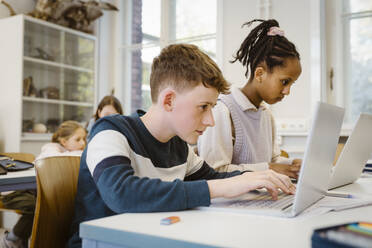 Focused schoolboy using laptop by female friend at desk in classroom - MASF38413