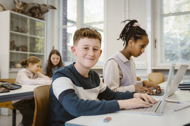 Porträt eines lächelnden Schülers mit Laptop bei einer Freundin im Klassenzimmer - MASF38414