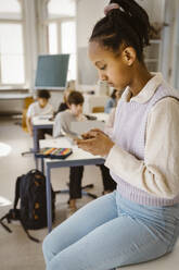 Schoolgirl surfing net through smart phone while sitting at desk in classroom - MASF38416