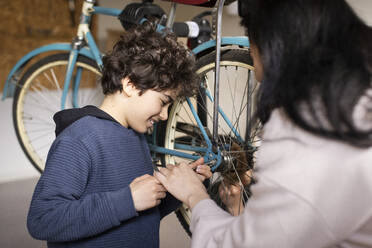 Smiling boy tightening screw of bicycle wheel with mother at recycling center - MASF38495