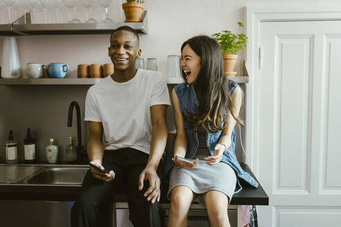 Cheerful young friends sitting with smart phones on kitchen counter at home - MASF38501