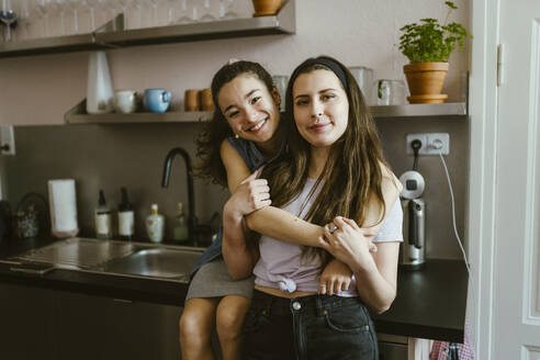 Portrait of happy woman hugging female friend from behind while sitting in kitchen at home - MASF38504