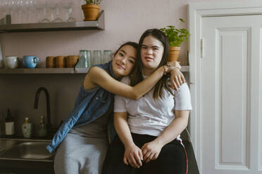 Junge Frau umarmt Schwester mit Down-Syndrom in der Küche zu Hause - MASF38510
