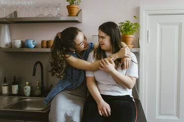 Smiling young woman hugging sister with down syndrome in kitchen at home - MASF38513