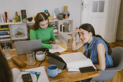 Smiling young woman sitting by friend studying at home - MASF38529