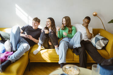 Happy young multiracial friends using smart phone while sitting together on sofa at home - MASF38539