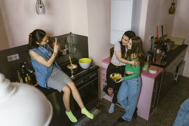 Smiling young woman sitting on counter photographing female friends in kitchen at home - MASF38555