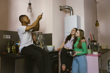 Smiling man sitting on counter photographing young friends in kitchen at home - MASF38556