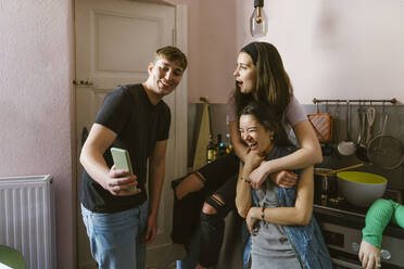 Smiling man sharing smart phone with cheerful female friends in kitchen at home - MASF38560