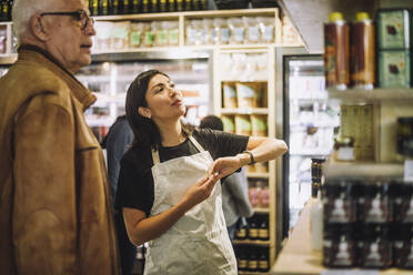 Saleswoman talking with senior male customer at delicatessen - MASF38604