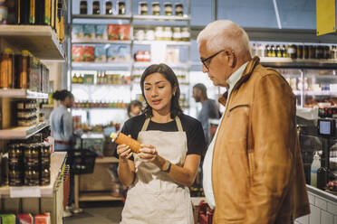 Saleswoman discussing over product with senior male customer at delicatessen - MASF38605