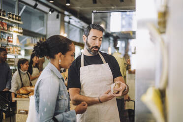 Mature salesman discussing over product with female customer at delicatessen - MASF38609