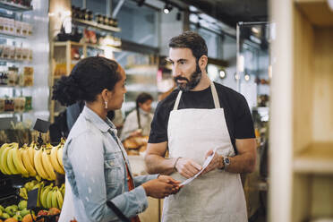 Mature salesman discussing over document with female customer at delicatessen - MASF38611