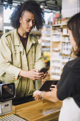 Young male customer scanning through smart phone while paying at checkout counter in store - MASF38616