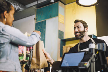 Smiling bearded retail clerk giving shopping bag to female customer at checkout - MASF38621