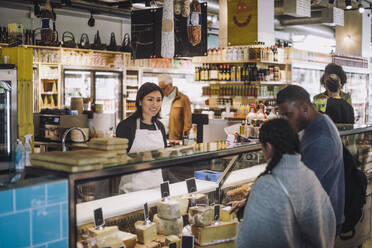 Smiling saleswoman looking at customers buying cheese from display cabinet at store - MASF38632