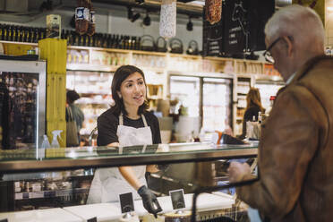 Saleswoman gesturing while talking with senior male customer at convenience store - MASF38633