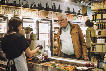 Saleswoman showing container to senior male customer at delicatessen - MASF38635