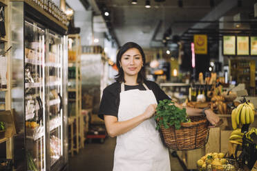 Porträt einer Einzelhandelsangestellten, die einen Weidenkorb trägt, während sie in einem Bio-Lebensmittelgeschäft steht - MASF38641